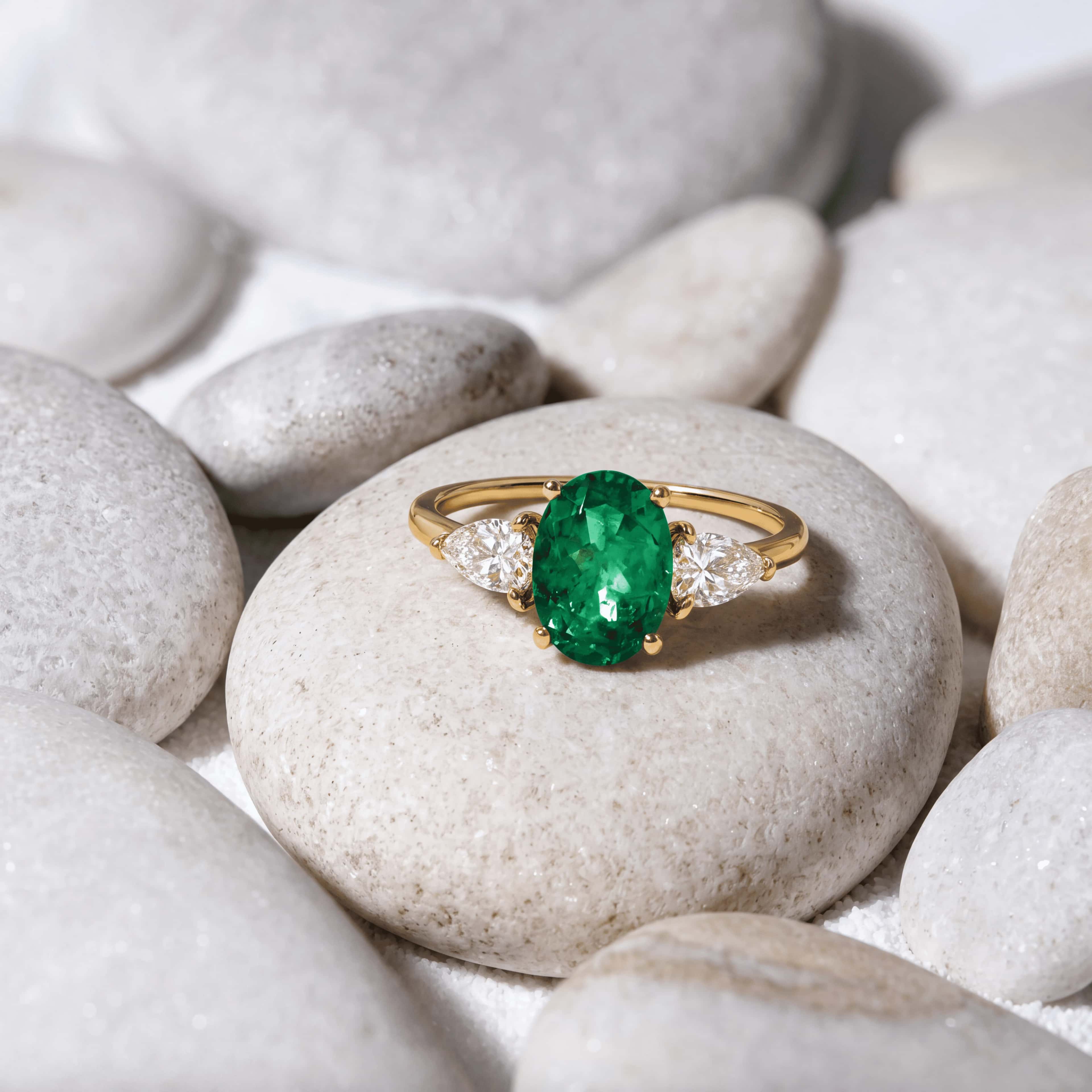 Oval emerald and pear diamond three-stone ring resting on pale pebbles