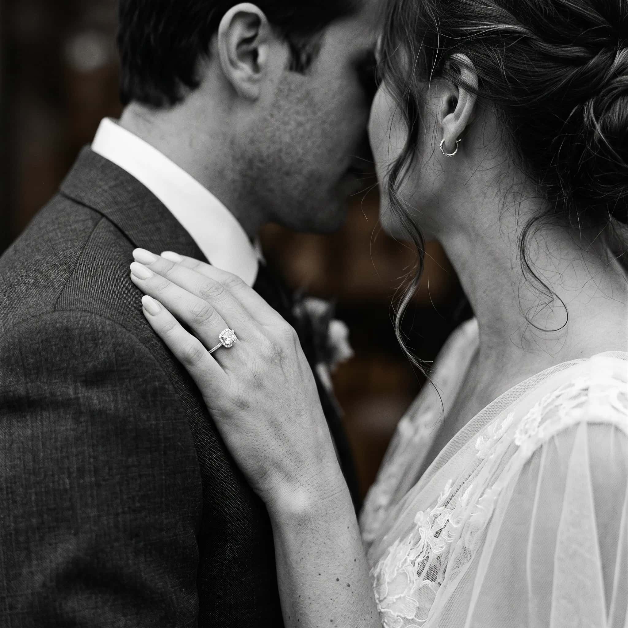 A couple embracing in wedding clothes, a cushion-cut halo engagement ring on the hand resting on a shoulder