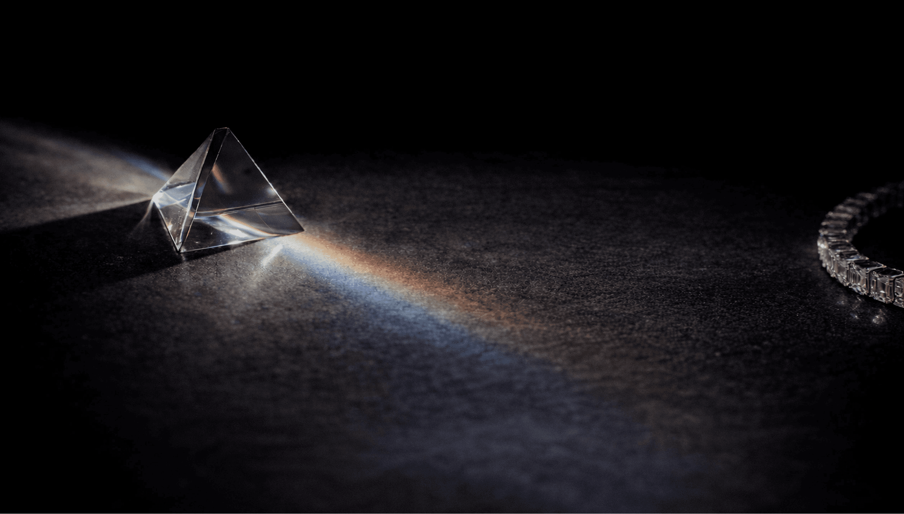 A wide shot of a glass prism with light shining through it, creating a spectrum of colors. Beside the prism, there is a pave bracelet made up of small diamonds, representing the brilliance and craftsmanship of MASAAT London.